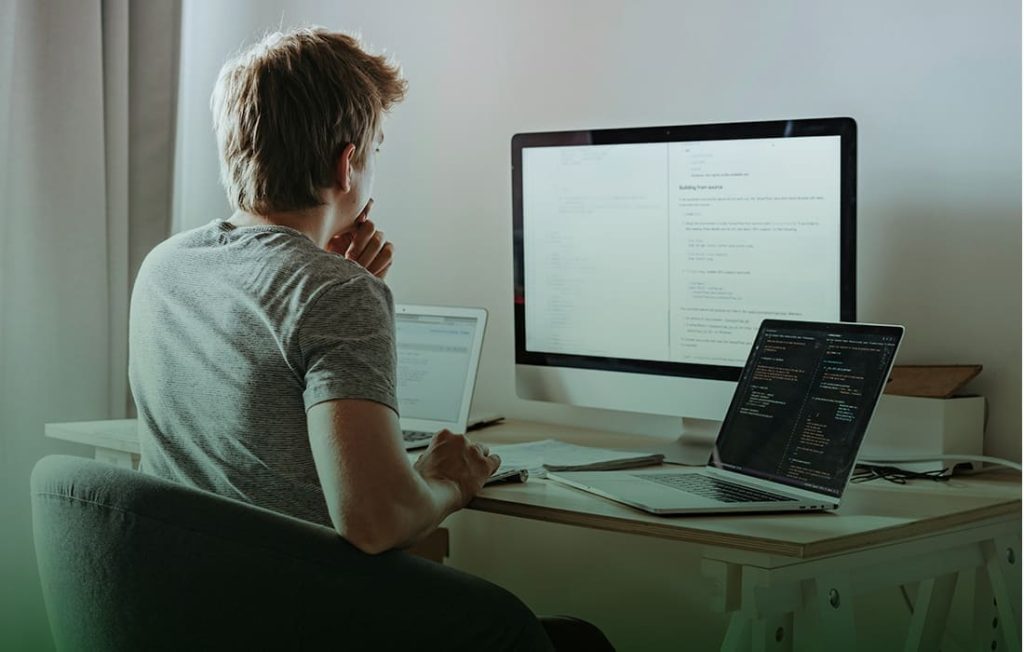 Person arbeitet am Schreibtisch mit mehreren Computern; grauer Stuhl, helle Wände, Bildschirme mit Text, ruhige Atmosphäre.